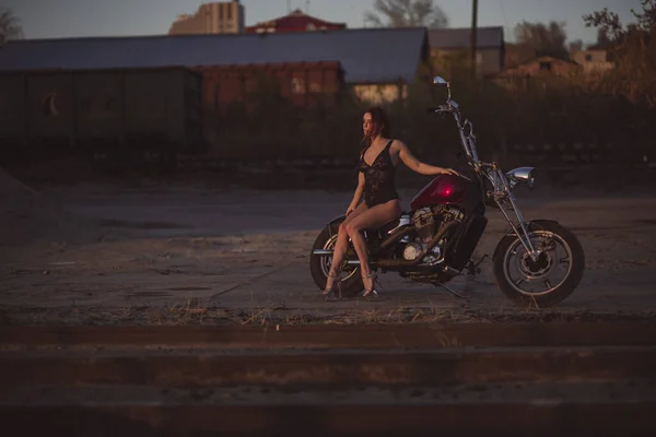Όμορφη σέξι γυναίκα σε δαντέλα bodysuit και ψηλά τακούνια κάθεται σε μια μοτοσικλέτα σε ένα βιομηχανικό φόντο — Φωτογραφία Αρχείου