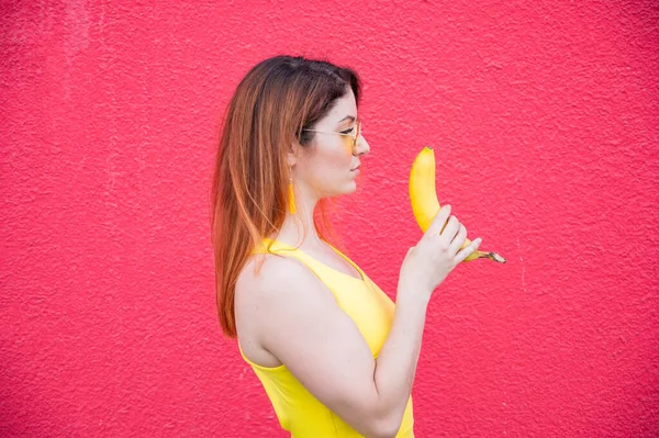 선글라스를 쓴 빨간 머리 여자가 빨간 배경에 바나나를 들고 있다 — 스톡 사진