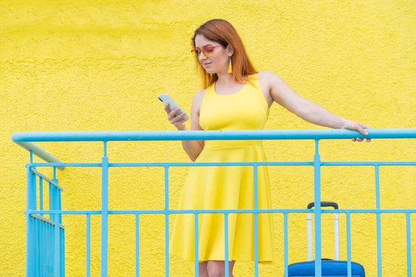 Krásná zrzavá žena stojí s modrým kufrem a používá smartphone na žlutém pozadí. — Stock fotografie