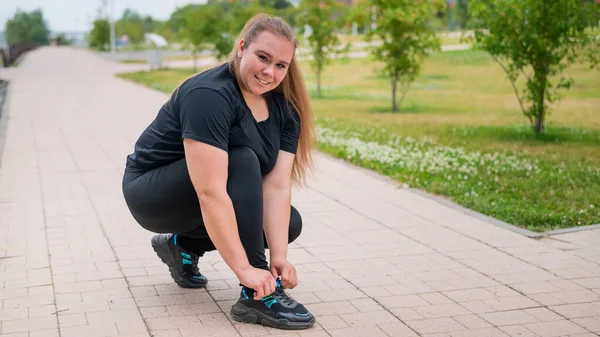 Una donna grassa in tuta si accovaccia e si lega i lacci delle scarpe all'aperto. — Foto Stock