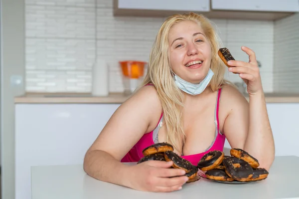 Een dikke blonde met grote borsten eet een donut met chocolade in de keuken. Een zwaarlijvige jonge vrouw heeft twee borden vol zoete gebak. Meisje met een medisch masker. — Stockfoto