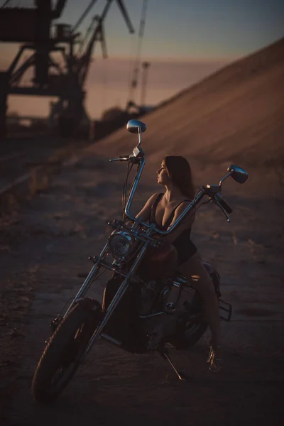 Beautiful sexy woman in lace bodysuit and high heels sits on a motorcycle on an industrial background — Stock Photo, Image