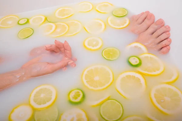 Mujer joven hermosa sin rostro toma un baño con limones de leche y lima. Fotografía recortada. Primer plano de las manos y pies femeninos en un baño de blanqueamiento de la piel. —  Fotos de Stock