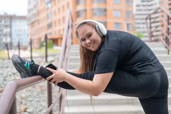 뚱뚱 한 젊은 여자가 스트레칭 연습을 하고 밖에서 헤드폰으로 음악을 듣는 모습 — 스톡 사진