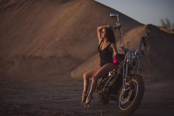 Hermosa mujer sexy en traje de encaje y tacones altos se sienta en una motocicleta en un fondo industrial — Foto de Stock