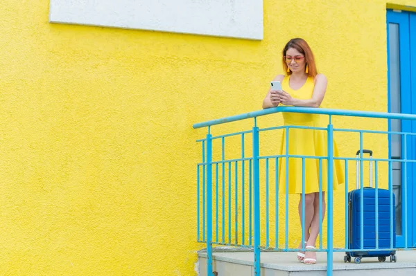 Bella donna dai capelli rossi si erge con una valigia blu e utilizza uno smartphone su uno sfondo giallo. — Foto Stock