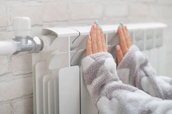 En kvinna i pyjamas värmer kalla händer på värmeelementet — Stockfoto