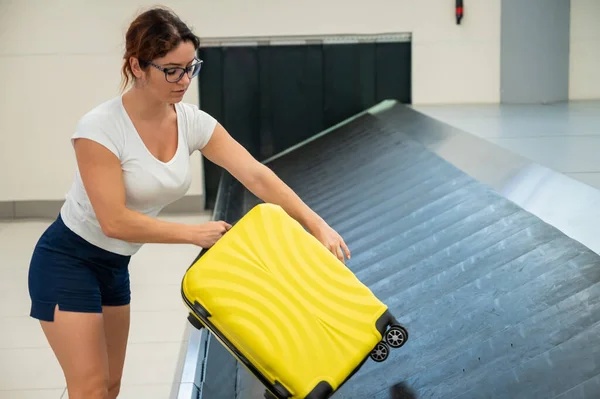 Mujer caucásica recoge su maleta amarilla del cinturón de equipaje en la terminal del aeropuerto. — Foto de Stock