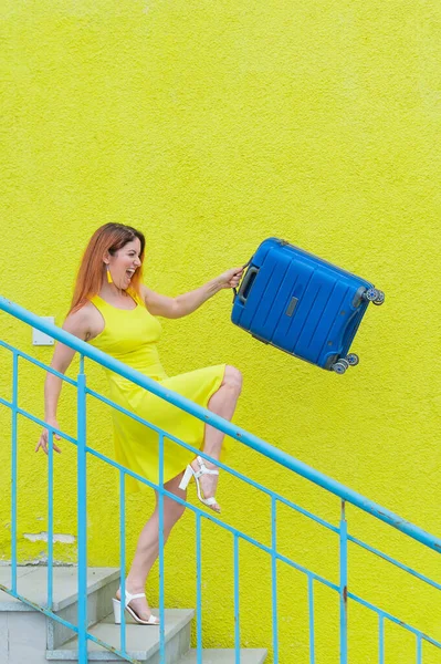 Šťastná zrzavá žena ve žlutých šatech jde nahoru a drží modrý kufr. — Stock fotografie