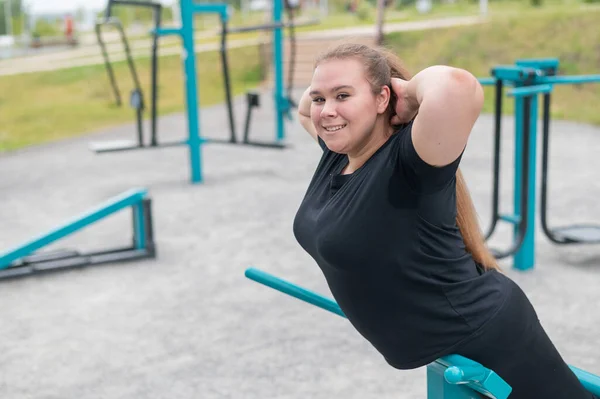 Tlustá mladá žena dělá prodloužení záda cvičení na cvičení stroje na sportovním hřišti venku. — Stock fotografie