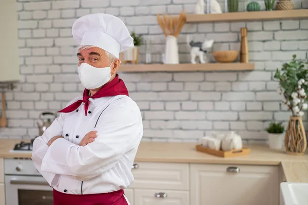 Un cuisinier âgé portant un masque dans la cuisine croise les bras sur la poitrine — Photo