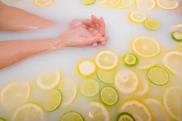 Belle jeune femme sans visage prend un bain avec des citrons au lait et de la chaux. Photo recadrée. Gros plan des mains et des pieds féminins dans un bain de blanchiment de la peau. — Photo