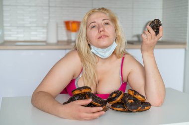 Koca memeli şişman bir sarışın mutfakta çikolata kaplı bir çörek yiyor. Obez bir genç kadın elinde iki tabak dolusu tatlı pasta tutuyor. Tıbbi maskeli kız..