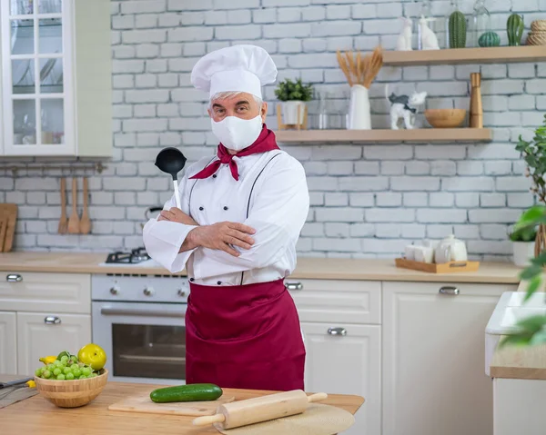 An elderly male chef wearing a mask in the kitchen crossed his arms over chest