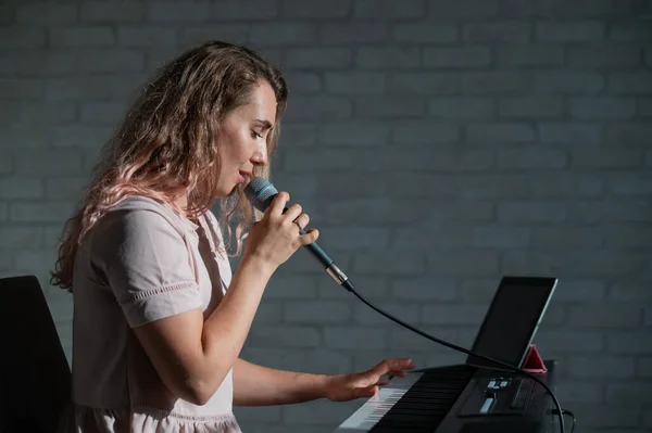 A woman records a vocal lesson using a laptop and accompanying on a keyboard while at home. The teacher sings a song into the microphone and plays the electronic piano. A blogger is recording a video.