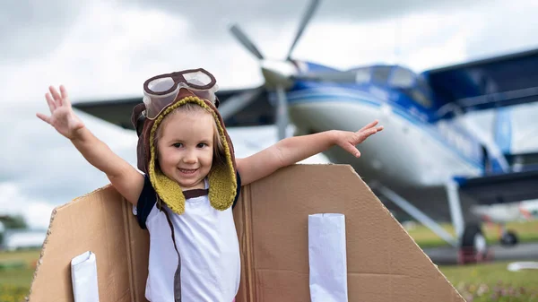 Ein kleines Mädchen im Pilotenkostüm mit Pappflügeln rennt vor der Kulisse des Flugzeugs über den Rasen. Ein Kind mit Hut und Brille träumt davon, im Flugzeug zu fliegen. — Stockfoto