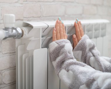 Pijama giyen bir kadın ısıtıcının üzerinde soğuk ellerini ısıtır.