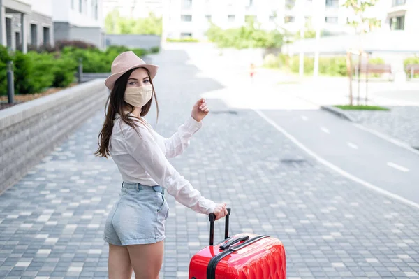 Een mooie jonge blanke vrouw met een medisch masker reist met een rode koffer door een lege stad. Het meisje gaat op vakantie met een grote bagage tas tijdens de coronavirus epidemie. — Stockfoto