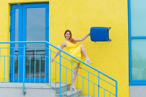 黄色のドレスを着た幸せな赤い髪の女性が階段を歩いていて、喜んで青いスーツケースを振っています。少女は旅の準備をしている。夏休みのコンセプト. — ストック写真