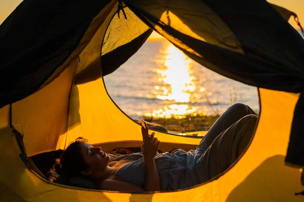 En kvinna ligger i ett turisttält vid floden och använder telefonen — Stockfoto