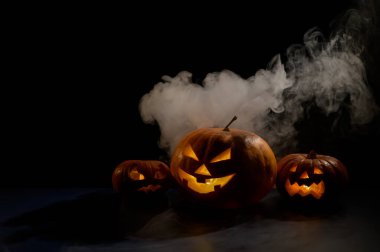 Siyah arka planda oyulmuş parlak bir sırıtışla üç ürkütücü cadılar bayramı buharı kabağı. El yapımı bir kriko-fener kafası ve içinde sis arasında karanlıkta bir mum. Şeker mi şaka mı? Kartpostal.