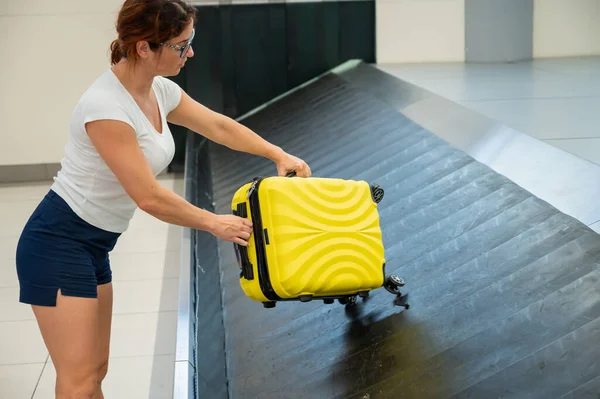 Mujer caucásica recoge su maleta amarilla del cinturón de equipaje en la terminal del aeropuerto. — Foto de Stock