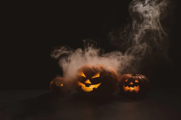 Three creepy halloween steaming pumpkins with a carved luminous smirk on a black background. A handmade jack-o-lantern head with a candle inside in the dark among the fog. Trick or treat. Postcard. — Stock Photo, Image