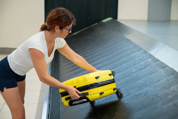 Mujer caucásica recoge su maleta amarilla del cinturón de equipaje en la terminal del aeropuerto. — Foto de Stock