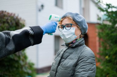 Doktor yaşlı bir kadının sıcaklığını ölçer. Maske takar ve açık havada kızıl ötesi termometre kullanmaz.