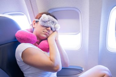 Kızıl saçlı yorgun bir kadın göz maskesi ve boyun yastığıyla uçakta uyuyor.