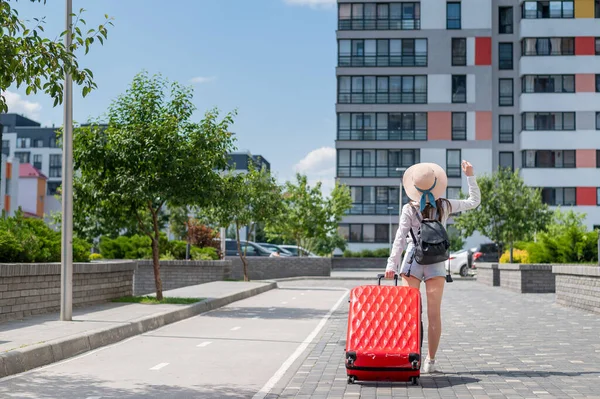 Schöne junge kaukasische Frau mit Sommermütze posiert mit einem roten Koffer im Freien. Glücklich lächelndes Mädchen geht mit einer großen Gepäcktasche in den Urlaub. Charmante Dame in kurzen Hosen an einem heißen, sonnigen Tag. — Stockfoto