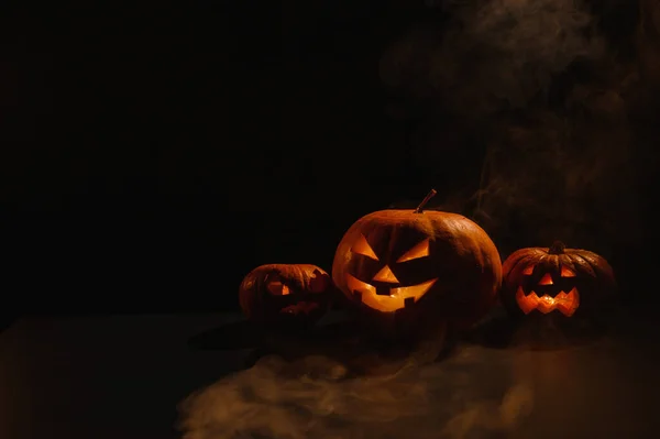Three creepy halloween steaming pumpkins with a carved luminous smirk on a black background. A handmade jack-o-lantern head with a candle inside in the dark among the fog. Trick or treat. Postcard. — Stock Photo, Image