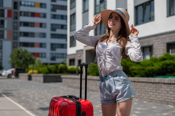 Mooie jonge blanke vrouw in een zomerhoed poserend met een rode koffer buiten. Gelukkig lachend meisje gaat op vakantie met een grote bagage tas. Charmante dame in korte broek op een hete zonnige dag. — Stockfoto