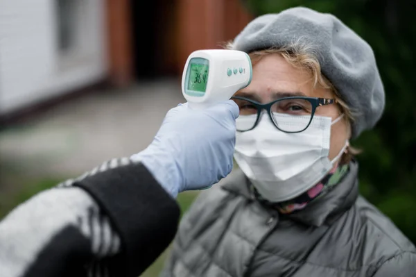 Un médico mide la temperatura de una mujer anciana que usa una máscara con un termómetro infrarrojo sin contacto al aire libre — Foto de Stock