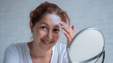 Portrait of a Caucasian woman with vitiligo uses sunscreen. A girl with a white pigment spot on her forehead looks in the mirror and is smeared with cream. clipart