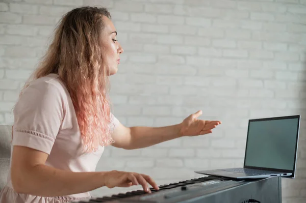 Uma mulher amigável toca piano eletrônico e conduz um vídeo blog em seu laptop. Fica em casa. Professor de instrumentos musicais. Música de aprendizagem à distância em quarentena. — Fotografia de Stock