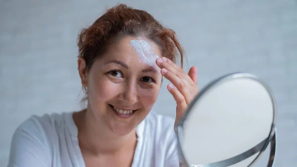 Porträtt av en vit kvinna med vitiligo använder solskyddsmedel. En flicka med en vit pigmentfläck i pannan ser sig i spegeln och är insmord med grädde. — Stockfoto