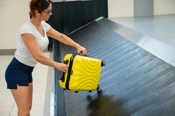 Mujer caucásica recoge su maleta amarilla del cinturón de equipaje en la terminal del aeropuerto. — Foto de Stock