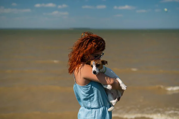 幸せな女性は海のそばの犬と抱擁します。ジャック・ラッセル・テリアの子犬を抱えている赤い髪の白人の少女。ペットとオーナーの愛. — ストック写真