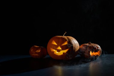 Cadılar Bayramı kartı. Siyah arka planda mumları parlayan bir fener. Bir sıra tüyler ürpertici balkabağı oyulmuş suratlar karanlıkta sigara içiyor..