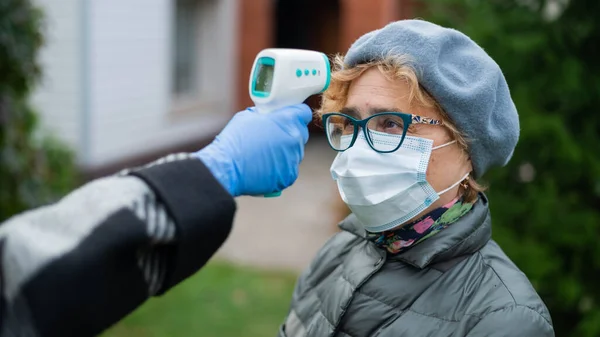 Un médico mide la temperatura de una mujer anciana que usa una máscara con un termómetro infrarrojo sin contacto al aire libre — Foto de Stock