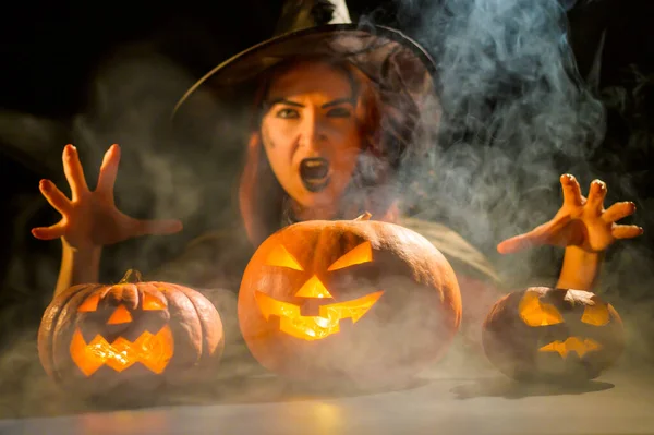 Złowroga wiedźma w kapeluszu przywołuje latarnię. Tradycyjne postacie halloween. Mistyczna mgła skrada się nad dyniami z wyrzeźbionymi okropnymi twarzami. Zła wiedźma rzuca zaklęcie. — Zdjęcie stockowe