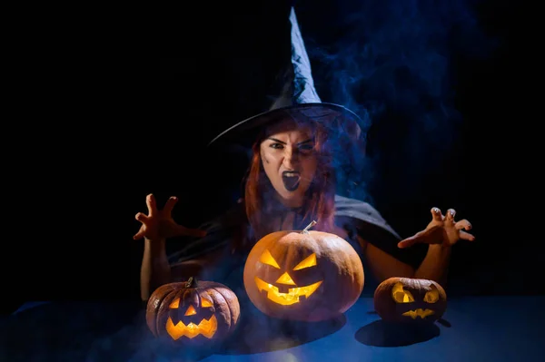 La malvada bruja lanza un hechizo sobre las calabazas. Retrato de una mujer en un disfraz de halloween de carnaval en la oscuridad — Foto de Stock