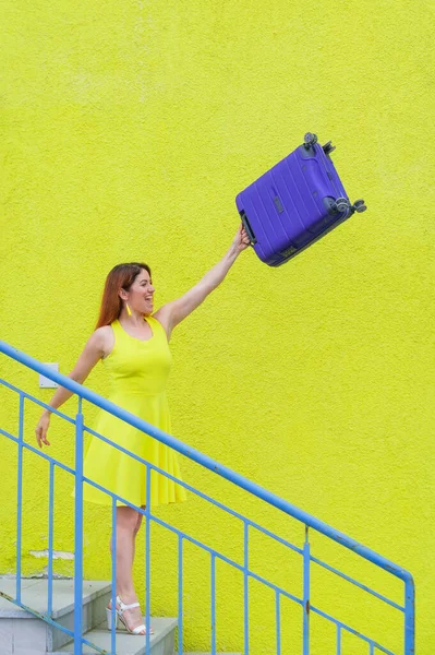 Krásná rusovlasá usměvavá žena radostně sestupuje po schodech a mává modrým kufříkem na žlutém pozadí — Stock fotografie