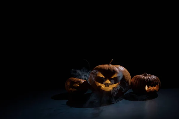 Tarjeta de Halloween. jack o linterna con velas brillan sobre un fondo negro. Una fila de calabazas espeluznantes con muecas talladas fuma en la oscuridad. — Foto de Stock