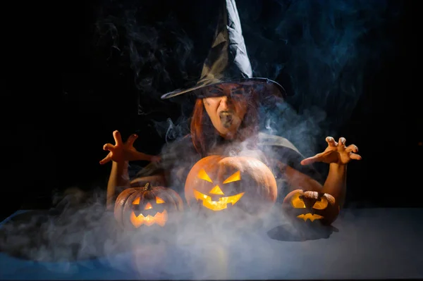 La malvada bruja lanza un hechizo sobre las calabazas. Retrato de una mujer en un disfraz de halloween de carnaval en la oscuridad — Foto de Stock