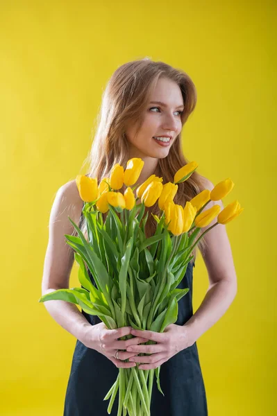 Kaukasische Frau mit einem Arm voller gelber Tulpen auf gelbem Hintergrund. Internationaler Frauentag. Strauß Frühlingsblumen — Stockfoto