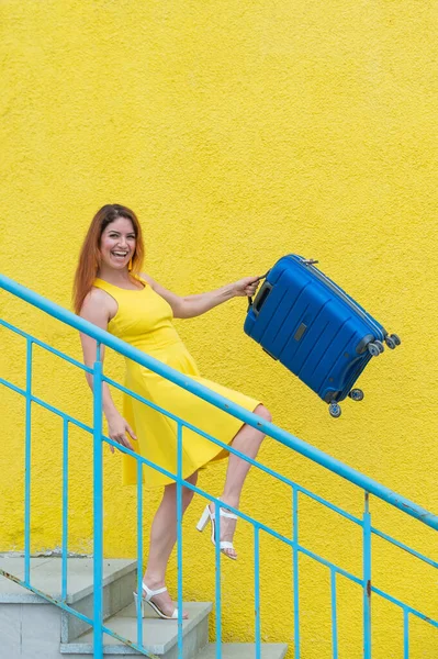 아름다운 빨간 머리웃는 여자가 기쁨스럽게 계단을 내려와 노란 배경에 파란 여행 가방을 흔들며 — 스톡 사진