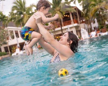 Beyaz bir kadın tatilde havuzda yüzerken oğlunu kucağına alıyor. Annesi küçük çocuğa yüzmeyi öğretiyor.