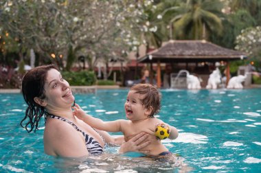 Beyaz bir kadın tatilde havuzda yüzerken oğlunu kucağına alıyor. Annesi küçük çocuğa yüzmeyi öğretiyor.